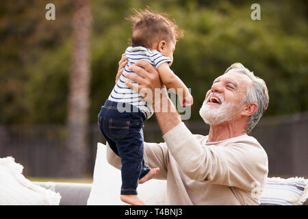 Senior Hispanic Mann im Garten sitzen heben sein baby Enkel in der Luft und zu ihm lächelnd Stockfoto