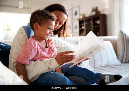 Nahaufnahme der jungen Mutter sitzt auf einem Sofa im Wohnzimmer ein Buch lesen mit Ihrem Kleinkind Sohn, der sitzt auf ihrem Knie, Seitenansicht Stockfoto