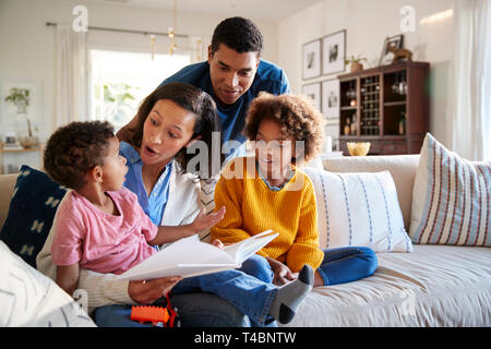 Junge Mutter sitzt auf einem Sofa im Wohnzimmer mit ihrer Tochter neben ihr und mein kleiner Sohn auf ihrem Knie, Vater steht hinter Ihnen, in der Nähe Stockfoto