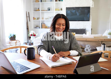 Happy mittleren Alters gemischten Rasse Frau am Tisch in Ihrem Esszimmer Notizen, selektiver Fokus sitzen Stockfoto
