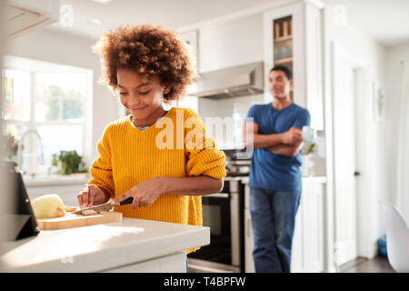 Vor - jugendlich Mädchen stehend auf der Arbeitsplatte in der Küche schnippeln, ihr Vater steht schiefen im Hintergrund lächelnd, selektiven Fokus Stockfoto