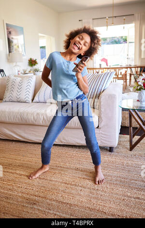 Vor - jugendlich schwarzes Mädchen singen und tanzen im Wohnzimmer, mit einem social media App auf Ihr Smartphone, volle Länge, vertikal Stockfoto