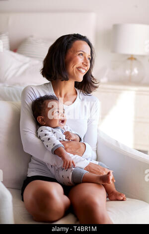 Junge Erwachsene Mutter sitzt in einem Sessel in Ihr Schlafzimmer, Ihr drei Monate alten Baby Sohn in ihre Arme und weg schauen lächelnd, vertikal Stockfoto