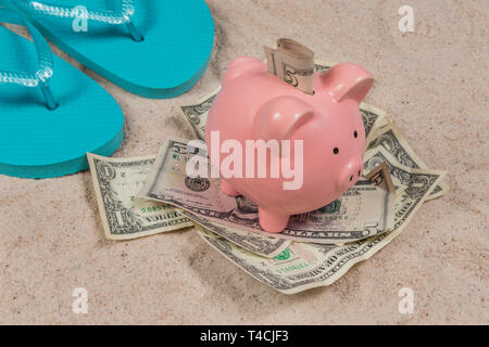 Horizontale Schuß von ein paar blaue flip-flops und ein rosa Sparschwein sitzt auf einem Haufen Geld mit Geld in den Steckplatz an einem Sandstrand. Stockfoto