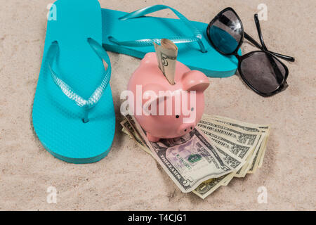 Horizontale Schuß von ein paar blaue flip-flops und ein rosa Sparschwein sitzt auf einem Haufen Geld mit Geld in den Steckplatz an einem Sandstrand Hintergrund. Es Stockfoto