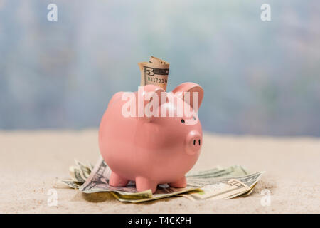 Horizontale Schuß eines rosa Sparschwein von der Seite sitzt auf einem Haufen Geld mit Geld in den Steckplatz an einem Sandstrand Hintergrund. Horizont nicht ganz ha Stockfoto