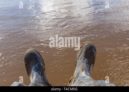 Füße eines Reisenden oder wilde Natur explorer in Schwarz schmutzigen Stiefel sitzt am Ufer des Flusses Stockfoto