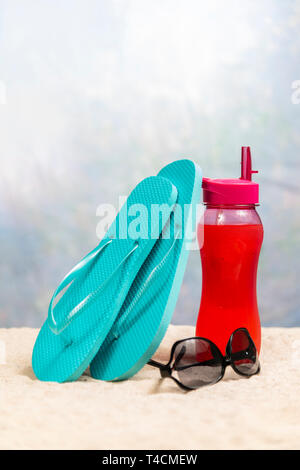 Vertikale geschossen von einem roten Getränk mit ein paar blaue flip-flops lehnte sich auf es und ein Paar schwarze Sonnenbrille zwischen ihnen. Alle auf einem sand Ferienhäuser beac Stockfoto