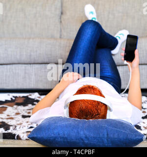 Ein nettes Mädchen in Kopfhörer verwendet ein Gadget. Liegen auf einer Decke. In der Nähe von dem Sofa zu Hause Stockfoto