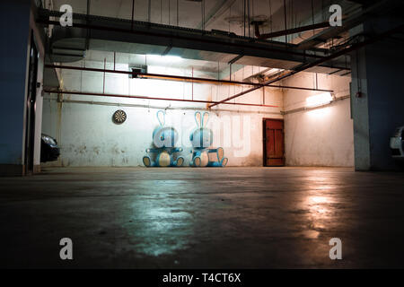 Zwei riesige blaue bunny Figuren auf dem Boden in einer dunklen, unheimlichen Tiefgarage, mit Grunge Wasserleitungen an der Decke und eine rostige Tür. Stockfoto