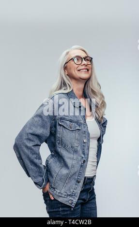 Stilvolle ältere Frau in Casuals bei copy space Blick auf grauen Hintergrund. Elegante Mitte nach kaukasische Frau in Denim Shirt und Brille suchen aw Stockfoto