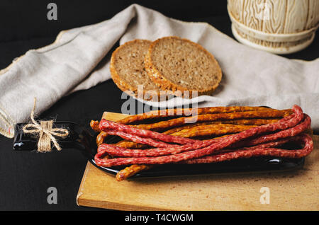 Lange dünne geräucherte Wurst und Brot auf schwarzen Hintergrund auf einer Platte von der Flasche. Traditionelle polnische Kabanosy oder Friesischen getrocknete Wurst st Stockfoto