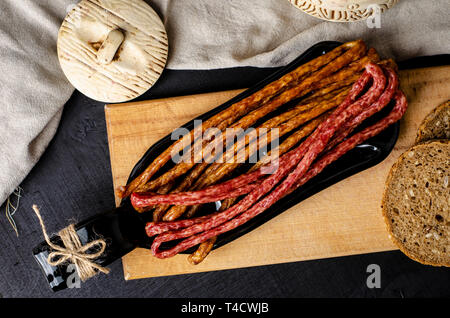 Lange dünne geräucherte Wurst und Brot auf schwarzen Hintergrund auf einer Platte von der Flasche. Traditionelle polnische Kabanosy oder Friesischen getrocknete Wurst st Stockfoto