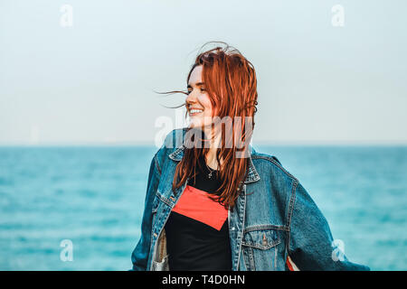 Portrait einer jungen Hübschen rothaarigen Mädchen in einer Jeansjacke im Hintergrund auf das Meer. Sie fühlt sich glücklich und frei, der Wind Wellen ihr Haar, sie suchen Stockfoto