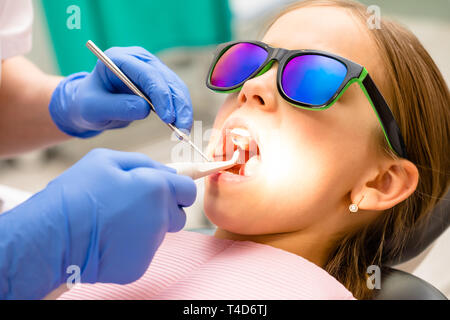 Zahnarzt untersucht die Zähne der elementare Alter Mädchen in pädiatrischen zahnmedizinischen Klinik mit Dental Werkzeug und gewinkelt Spiegel. Kind in einem behandlungsstuhl Sitzen tragen Stockfoto