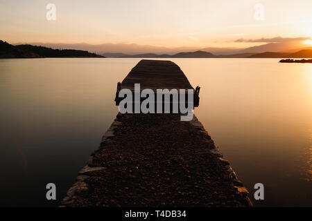 Sonnenaufgang über eine kleine hölzerne Pier in ruhigem Wasser mit gelben Töne, aufgehende Sonne und majestätischen Bergen (Kalami, Korfu, Griechenland, Europa) Stockfoto