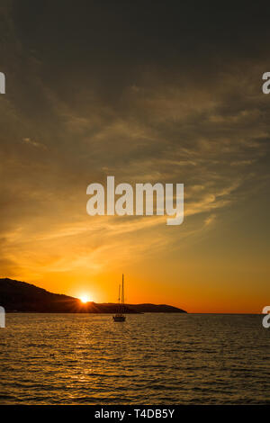 Dramatische rot und orange Sonnenuntergang über dem Meer mit Gebirge im Hintergrund und Yacht auf dem Wasser (Kassiopi, Korfu, Griechenland, Europa) Stockfoto