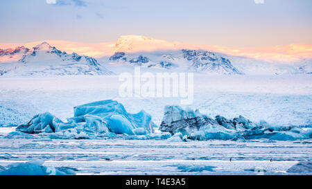 Eisberge schwimmen auf Gletschersee Jökulsárlón Gletscherlagune bei Sonnenaufgang, mit Hintergrund Berggipfel von Sunrise beleuchtet, in Island. Stockfoto