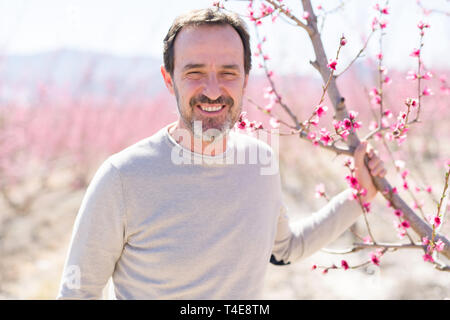 Stattliche mittleren Alters älterer Mann lächelt fröhlich auf einem Pfirsich- Garten mit rosa Blütenblätter an einem sonnigen Tag der Feder Stockfoto