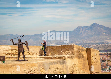 Frau Touristen auf der Suche nach Telefon vor Stahl Skulptur eines Mittelalterlichen Soldat warf einen Speer in Castell de la Santa Barbara, Alicante Spanien Stockfoto