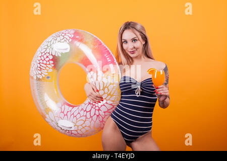 Glückliche junge Frau im Badeanzug holding Sommer Cocktail auf gelben Hintergrund. Haltering schwimmen. Attraktive Frau. Fröhliche Frau. Stockfoto