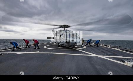 Ozean (7. April 2019) Matrosen an Bord der Arleigh-Burke-Klasse geführte Anti-raketen-Zerstörer USS Carney (DDG64) die Keile und Ketten von einem MH-60S Seahawk, zu der "Ritter" der Hubschrauber Meer Combat Squadron (HSC) 22, während der Übung gemeinsame Krieger 19-1, 7. April 2019 vergeben entfernen. Gemeinsame Krieger ist ein Vereinigtes Königreich - led, multinationale Übung in einem robusten maritime Ausbildung Umfeld, in dem die Verbündeten und Partnern Interoperabilität verbessern kann und Kräfte für kombinierte Operationen vorzubereiten. Stockfoto