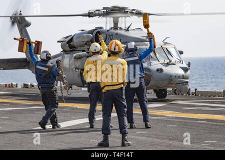 Straße von Hormuz (April 3, 2019) - der Luftfahrt Bootsmann Gehilfen (Handling) vorhanden Keilen und Ketten der Pilot eines MH-60S Sea Hawk Hubschrauber, die sich anschickt, aus dem Flight Deck der Wasp-Klasse amphibisches Schiff USS Kearsarge (LHD3) während der Durchfahrt der Straße von Hormuz. Kearsarge ist das Flaggschiff der Kearsarge amphibische Gruppe und bereit, mit der begonnen 22 Marine Expeditionary Unit, ist in die USA 5 Flotte Bereich der Maßnahmen zur Unterstützung der Marine im Einsatz für die Stabilität und Sicherheit in der Region zu gewährleisten und verbindet das Mittelmeer und die Stockfoto