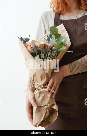 Frau in eine braune Schürze mit roten Haaren halten Schönen Blumenstrauß von lebenden Korallen farbigen Rosen und eryngium auf einem Licht Stockfoto