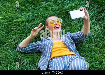 Glückliche junge kurzes Haar Frau in casual blau gestreiften Anzug, gelbes Hemd, Gläser liegend auf grünem Gras mit Handy, tut selfie oder online vi. Stockfoto