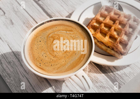 Frühstück im Sonnenlicht mit Vintage Holz- Hintergrund, weiße Porzellan Tasse mit heißem aromatischen Kaffee mit Schaum und Wiener Waffeln auf einem Teller. Stockfoto