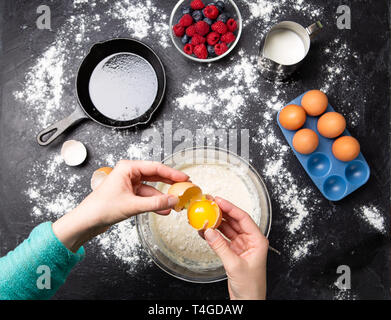 Foto der Hände des Menschen Aufschlagen der Eier in der Schale. Tabelle mit Beeren, Milch, Mehl. Stockfoto