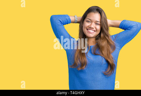 Junge schöne Brünette Frau trägt blaue Pullover über isolierte Hintergrund Entspannung und Stretching mit Armen und Händen hinter dem Kopf und Hals, lächelnd Stockfoto