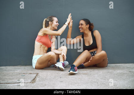 Zwei passen junge Frauen in Sportkleidung auf dem Boden vor Lachen und hohe Fiving, nachdem Sie arbeiten zusammen Stockfoto