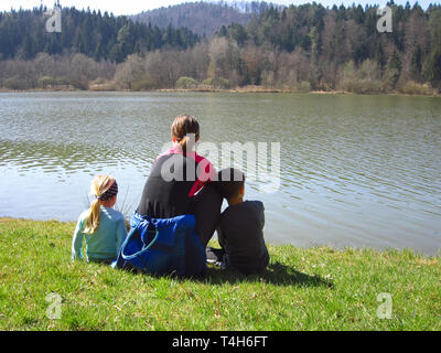 Mutter mit Sohn und Tochter sitzen auf einer Wiese des grünen Grases am See und Wald. Glückliche Familie, in der Natur Stockfoto
