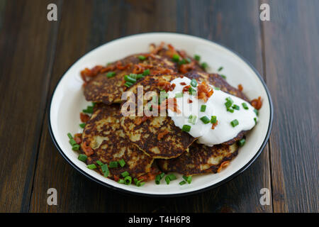 Kartoffelpuffer draniki serviert mit Sauerrahm, Schnittlauch und Speck auf einem weißen Teller. Dunkler Holztisch, Blau Leinen Servietten, hohe Auflösung Stockfoto