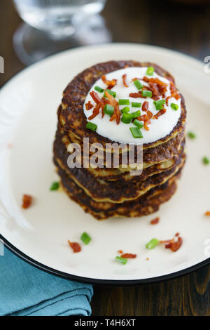 Kartoffelpuffer draniki serviert mit Sauerrahm, Schnittlauch und Speck auf einem weißen Teller. Dunkler Holztisch, Blau Leinen Servietten, hohe Auflösung Stockfoto