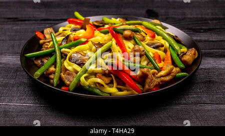 Leckere gebratene Nudeln mit Knoblauch Sprößlinge, Pilze, Paprika und Hähnchenfleisch auf einem schwarzen Hintergrund einer rustikalen Holztisch rühren. Eine beliebte Stockfoto