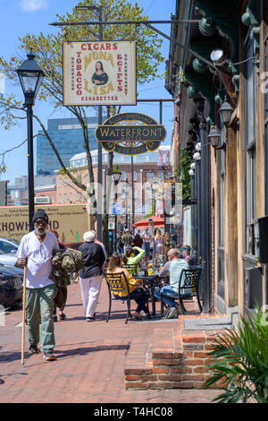 Fells Point, Baltimore, MD, USA - 13. April 2019. Ein Mann mit einem Stock die Spaziergänge entlang der Themse St während andere Frühstück in einem Café im Freien in Fells Poi haben Stockfoto