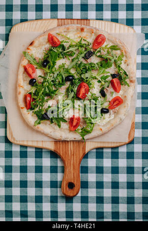 Neapolitanische Pizza auf Sahnesoße mit Thunfisch, Käse, Rucola, Basilikum, Tomaten, Oliven, bestreut mit Käse auf einem Holztisch auf einer Tischdecke in einer Zelle Stockfoto