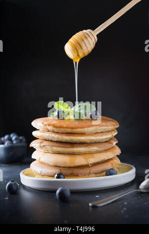 Süße hausgemachte Stapel Pfannkuchen mit frischen Heidelbeeren, Honig und Minze in der modernen Platte auf schwarzen Hintergrund. Stockfoto