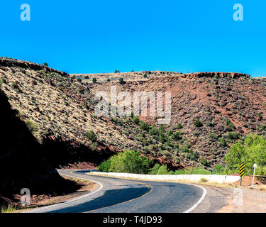 Tow Lane Country Road durch Red Hills mit grünen und gelben Pinsel geschwungen unter einem blauen Himmel. Stockfoto