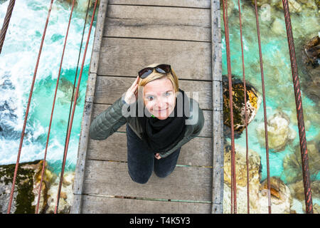 Glücklich lächelnde und entspannende Frau auf einem Tagesausflug auf eine alte hölzerne Hängebrücke über die fließende kristallklare türkisfarbene Fluss Soca, Slowenien Stockfoto