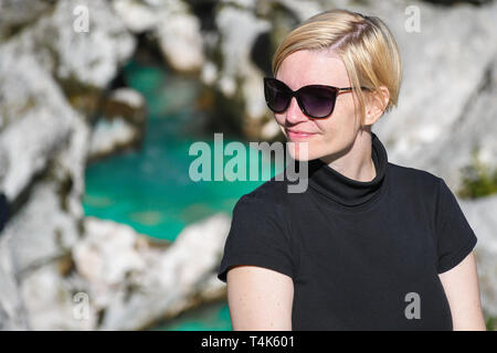 Glücklich lächelnde Frau mit schwarzer Sonnenbrille und t-shirt neben einem schönen türkisfarbenen Soca River Canyon im Hintergrund posiert, Slowenien Stockfoto