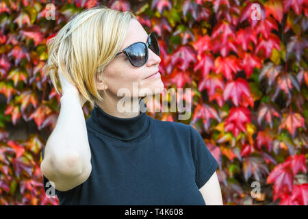 Gerne entspannt Frau mit schwarzer Sonnenbrille und t-shirt Halten Sie ihren Arm auf der Rückseite des Kopfes und neben einem schönen Herbst farbige posing Wand Blätter Stockfoto