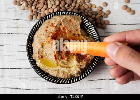 Hohe Betrachtungswinkel eines Mannes Eintauchen ein Streifen von Karotte in einem hausgemachten Linse hummus gewürzt mit Paprika in einer grünen Keramik Teller serviert, auf einem weißen Tropfen Stockfoto