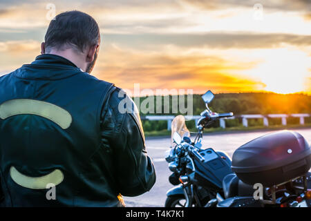 Biker in eine Lederjacke Neben dem Motorrad auf dem Hintergrund bei Sonnenuntergang stehend Stockfoto