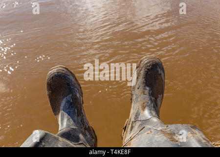 Beine von einem Fischer in schmutzigen Gummistiefel sitzt am Ufer des Flusses mit schmutzigem Wasser im Hintergrund Stockfoto