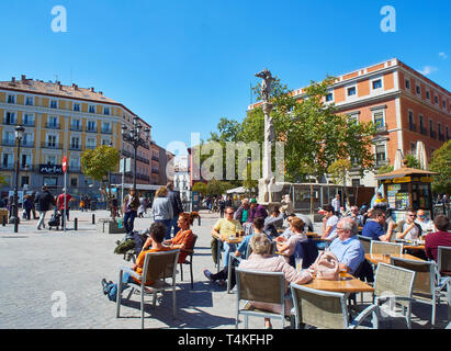 Leute erfrischend auf einer Terrasse im Jacinto Benavente Square von Madrid mit der Jakobsweg Kreuzung Stein im Hintergrund. Stockfoto