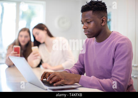 Teenager mit Laptop zu Hause, während Mädchen an Handys im Hintergrund Stockfoto