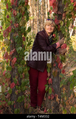 Eine glückliche Frau mittleren Alters ist im Wald im frühen Herbst. Rosa Kleidung. Es steht in der Nähe von schönen Bäumen. Es gibt große rosa Blätter um. Stockfoto